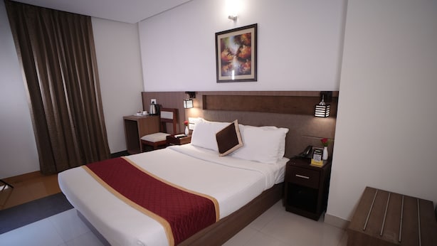 A hotel room with a large bed, a nightstand, a desk, a chair, a lamp, and a painting on the wall. - HOTEL DE GRAND MALABAR (Kozhikode)