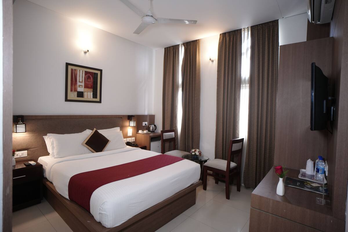 A hotel room with a large bed, bedside tables, a desk, a chair, a TV, and a window with curtains.