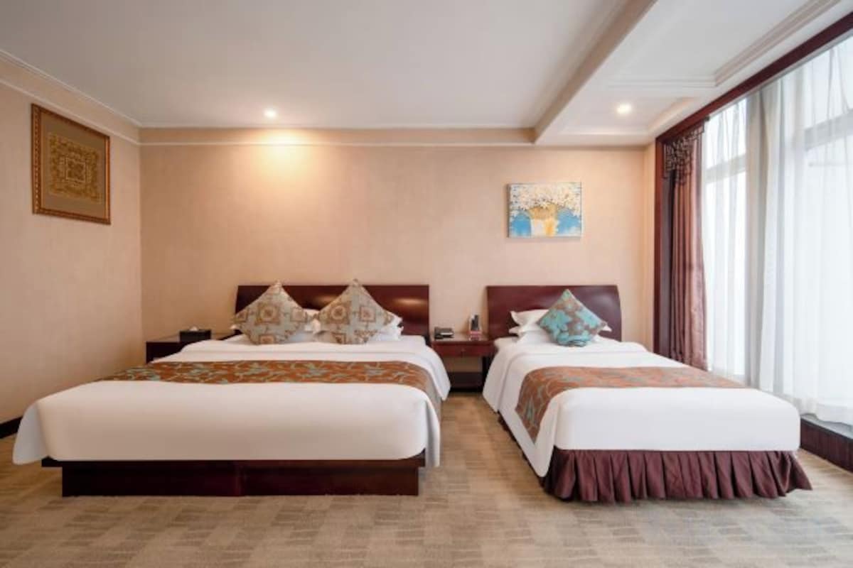 A hotel room with two beds, a nightstand, a painting on the wall, and a large window with curtains.