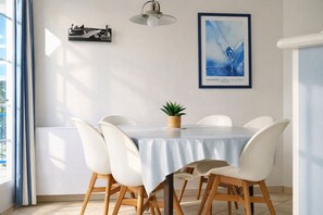 A dining area with a round table, white chairs, a light blue tablecloth, a hanging light fixture, a framed picture on the wall, and a small potted plant.