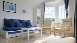 A living room with a blue sofa, a white coffee table, two wooden armchairs, and a view of the outdoors through large windows.