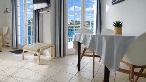 A room with a bed, a table with a tablecloth, chairs, a TV mounted on the wall, and large windows with curtains.