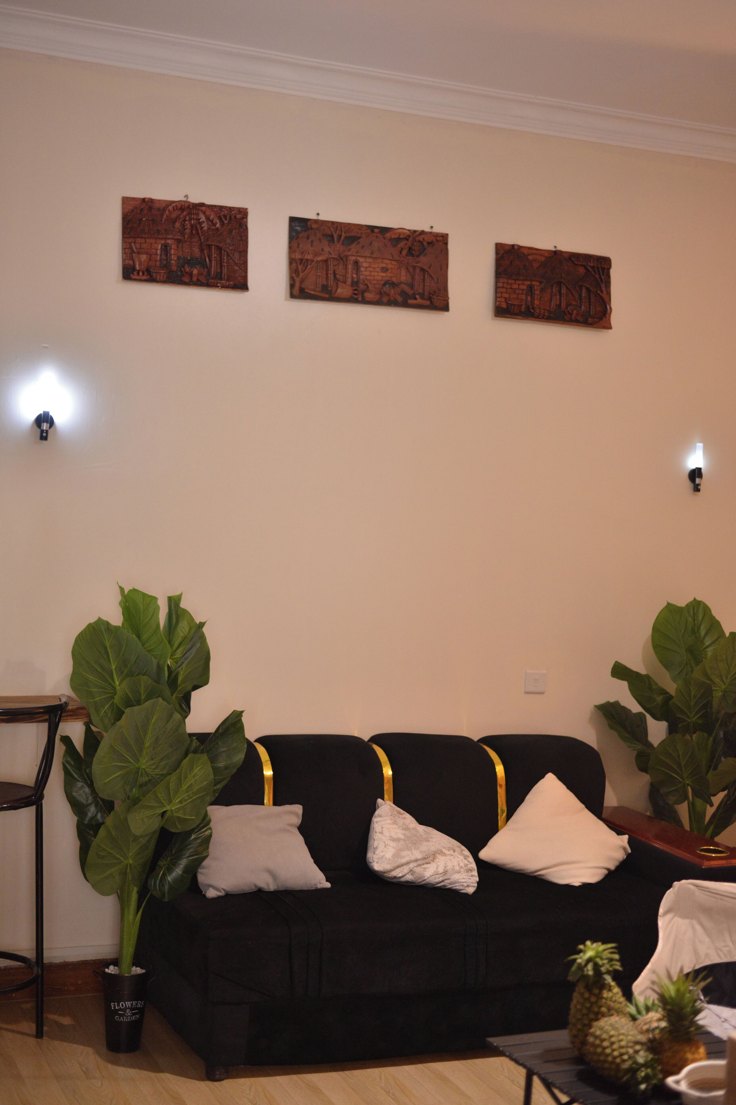 A living room with a black sofa, two large potted plants, and three wall-mounted artworks.
