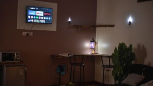 Il s’agit d’un espace de vie compact doté d’un téléviseur à écran plat fixé au mur, qui diffuse différents services de streaming. On y trouve une petite kitchenette équipée d’un four à micro-ondes et d’un réfrigérateur. Un bureau avec deux chaises est placé près du téléviseur, et une plante en pot est visible au premier plan.