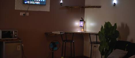 A compact living space with a flat-screen TV mounted on the wall displaying various streaming services. There is a small kitchenette area with a microwave and a refrigerator. A desk with two chairs is positioned near the TV, and a potted plant is visible in the foreground.