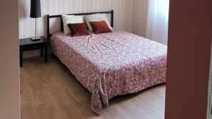 Een slaapkamer met een houten vloer, een bed met een beddegoed met patroon, een nachtkastje met een lamp, een aan de muur hangend schilderij en een plafondlamp.