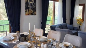 A dining area with a long table set for four, featuring white plates, gold cutlery, and candles. There is a painting on the wall and a sofa in the background.