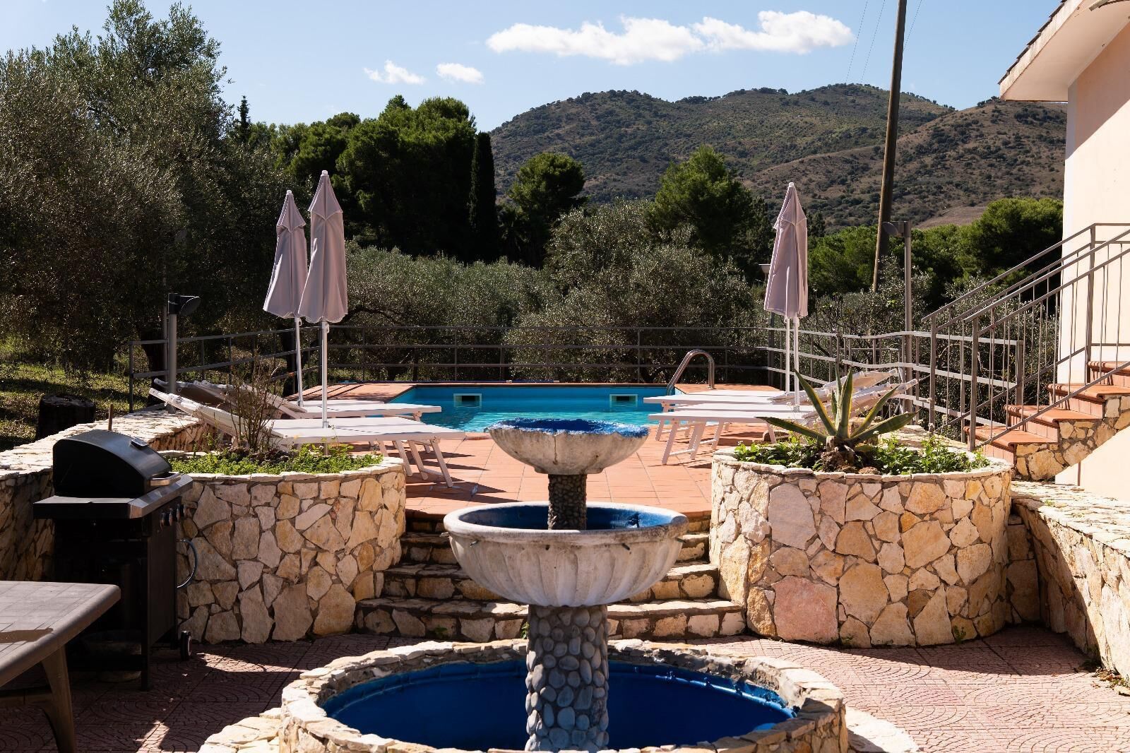 A pool area with a fountain, stone planters, lounge chairs, and a grill.