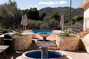 A pool area with a fountain, stone planters, lounge chairs, and a grill.