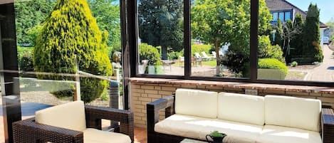 A sunlit patio with wicker furniture and a glass table, overlooking a garden with a tall tree and a house in the background.