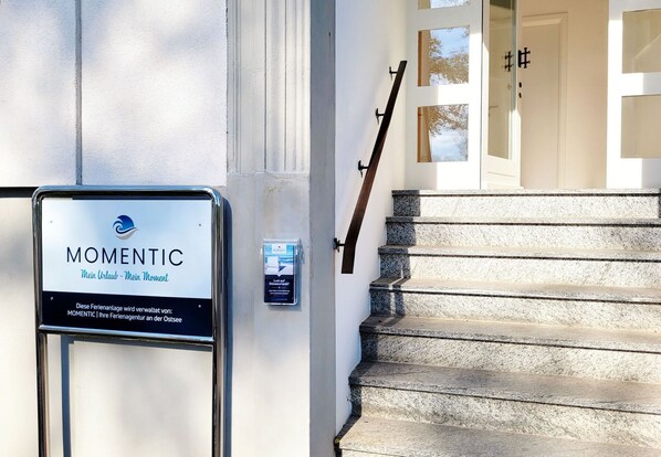 Placa de 'MOMENTIC' com logotipo, escadas e uma porta com placa de número.