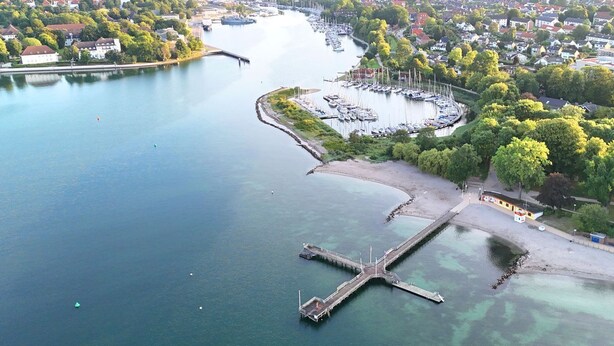 An aerial view of a riverside town with a marina, a bridge, and residential areas. - Charming apartment by the harbor with free bed linen package and terrace - (Neustadt in Holstein)