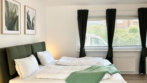 Ein ordentlich bezogenes Bett mit einer grünen Tagesdecke, einem grünen, mit Stoff bezogenen Kopfteil und weißer Bettwäsche. An der Wand befinden sich zwei gerahmte Pflanzenbilder.
