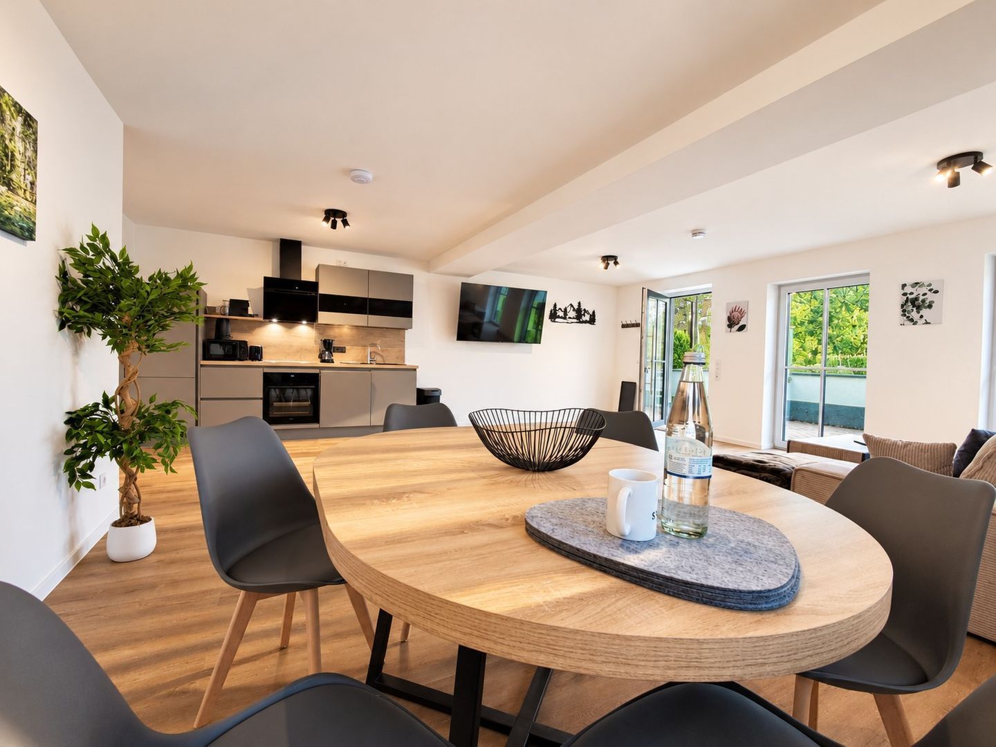 A modern kitchen with built-in appliances, a dining area with a round table, and a large window providing a view of greenery.