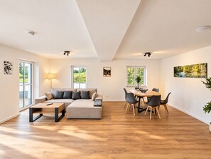 Ein großzügiger Wohnbereich mit Sofa, Esstisch, Stühlen, großen Fenstern und einer Decke mit eingelassener Beleuchtung.