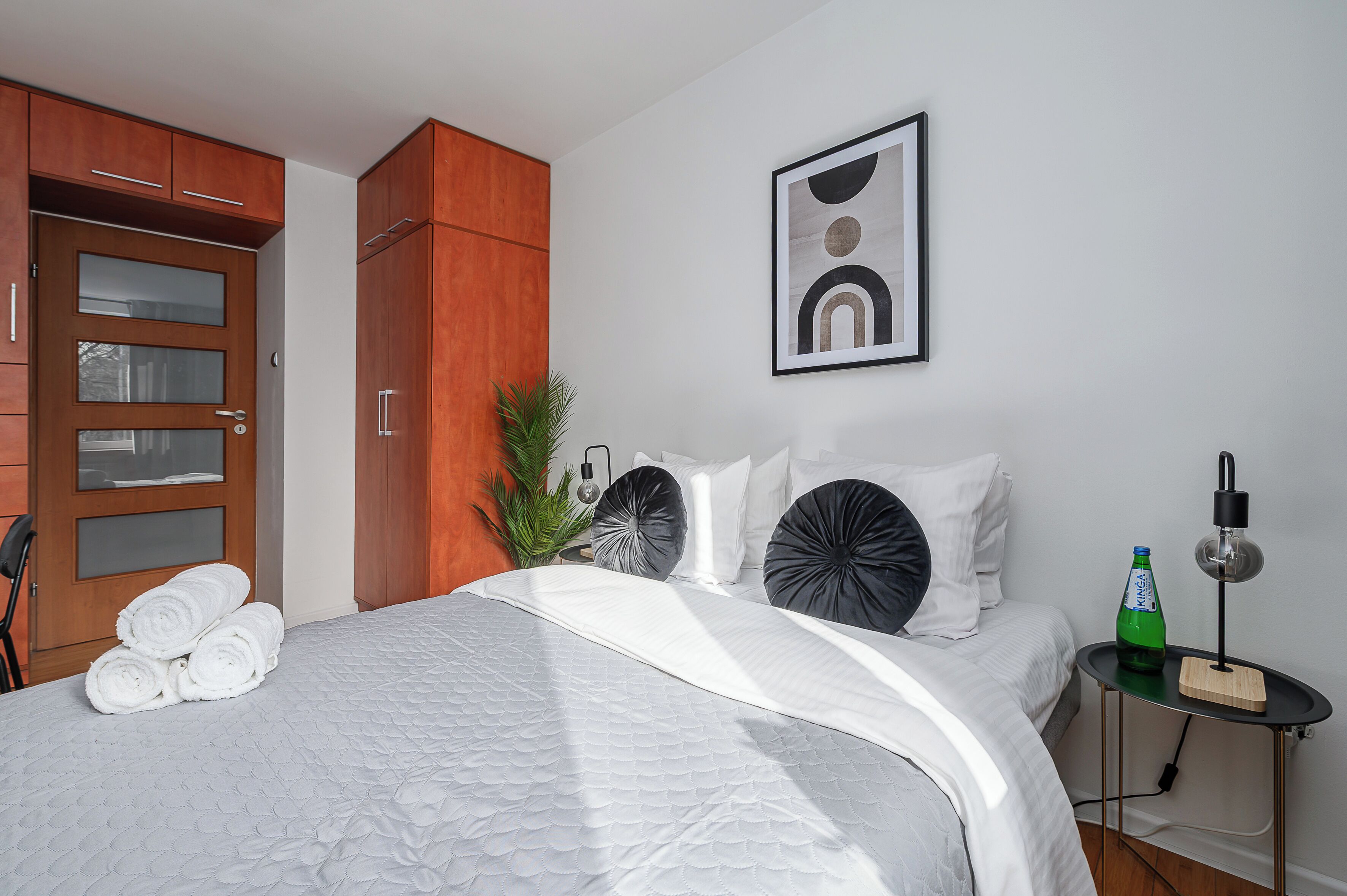 Habitación de hotel con cama, armario de madera, cuadro abstracto enmarco, botella verde y lámpara de escritorio.