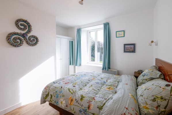 A bedroom with a floral bedspread, a window with curtains, a wall-mounted picture, and a door.