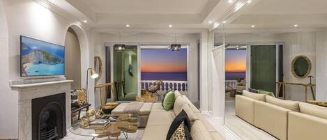 A spacious living room with a fireplace, a large sofa, a television, and a view of the sea through a glass door.