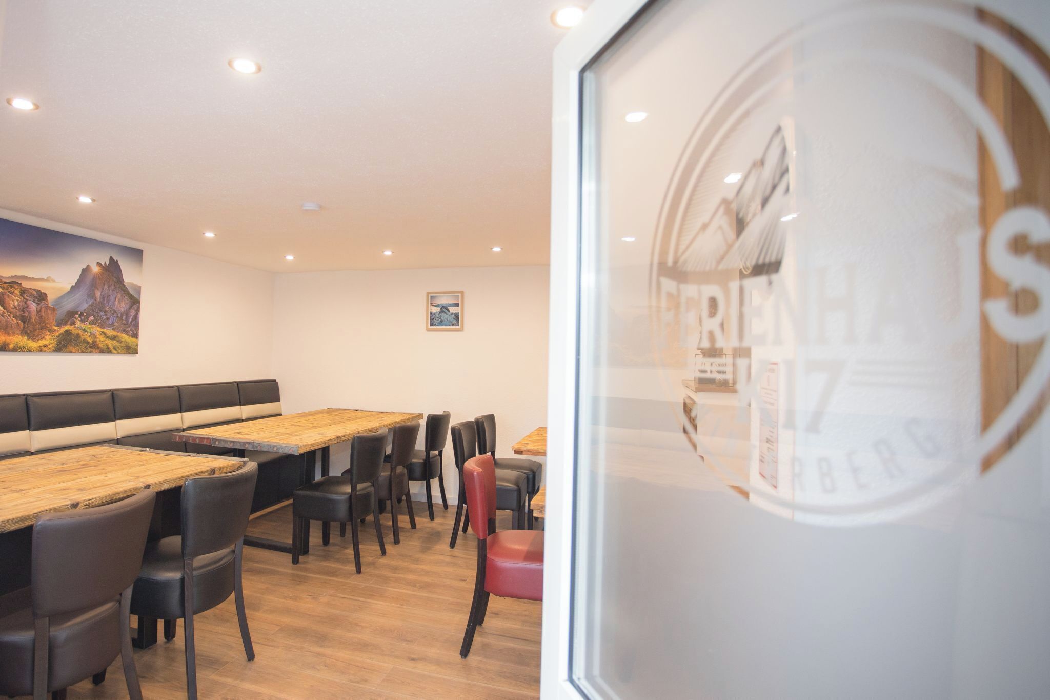 Een restaurantinterieur met houten tafels, zwarte en rode stoelen en een schilderij van een berglandschap aan de muur.