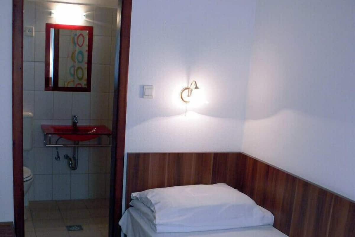 A small, single-bed room with a wooden headboard, a red-framed mirror, a white sink, and a wall-mounted light fixture.
