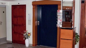Ein Empfangsbereich mit einer Rezeption, einem blauen Aufzug, einer roten Tür mit der Aufschrift 'TOILET' und einer weißen Vase mit Blumen.