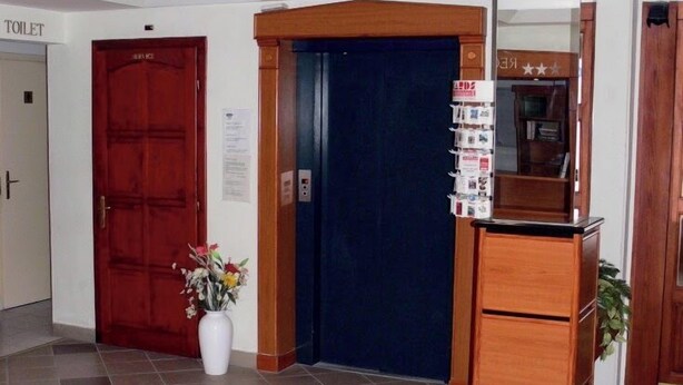 A reception area with a reception desk, a blue elevator, a red door labeled 'TOILET,' and a white vase with flowers. - Hid Hotel (Budapest)