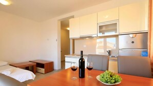 Una cocina moderna con armarios blancos, microondas integrado y refrigerador de acero inoxidable. Hay una mesa de comedor de madera con una botella de vino y copas, y un bol con uvas.