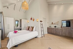 A spacious bedroom with a large bed, a wooden ladder, a kitchenette, and a ceiling with exposed beams.