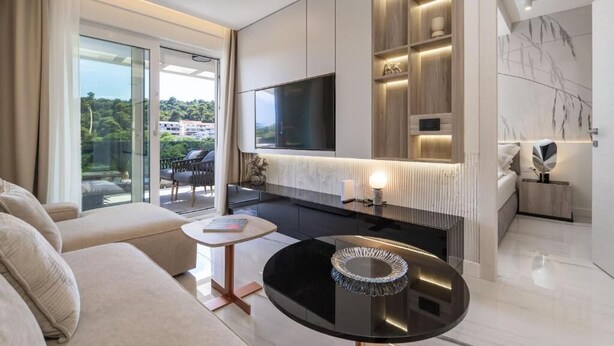 Een moderne hotelkamer met een grote televisie, een balkon met buitenmeubilair en een strakke, zwarte salontafel.