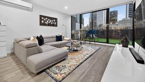 A modern living room with a grey sectional sofa, a coffee table, a patterned rug, and a large window with a city view. - Spacious 3BR on Bourke St Grand Balcony & City Views (Melbourne)
