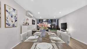 A modern dining room with a wooden table set for four, a vase of flowers, and a television on the right. - Spacious 3BR on Bourke St Grand Balcony & City Views (Melbourne)