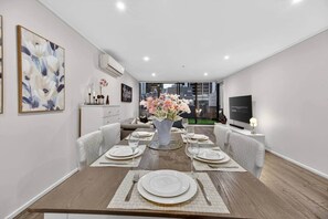 A modern dining room with a wooden table set for four, a vase of flowers, and a television on the right. - Spacious 3BR on Bourke St Grand Balcony & City Views (Melbourne)