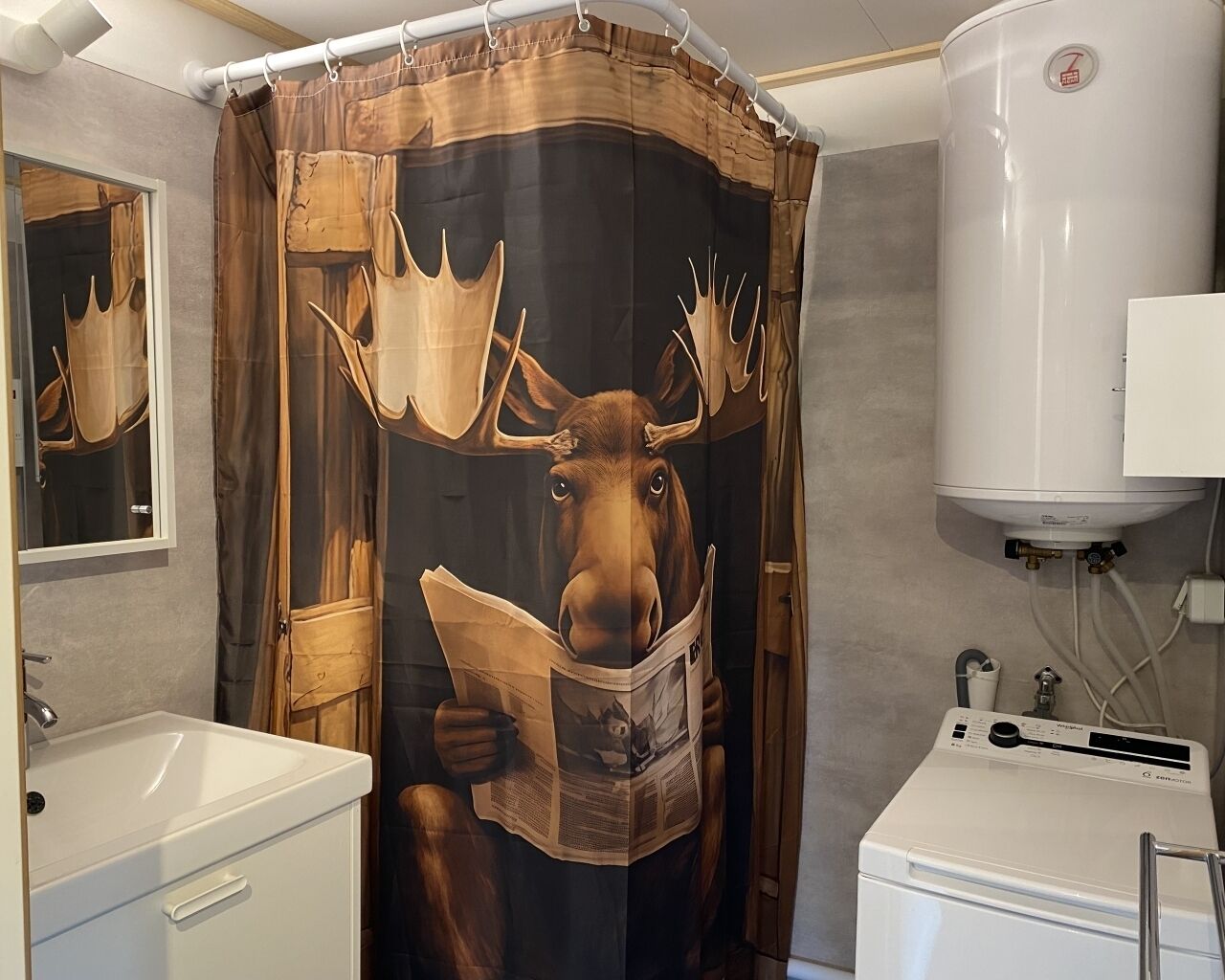 A bathroom with a shower curtain featuring a moose reading a newspaper, a white sink, and a white toilet.