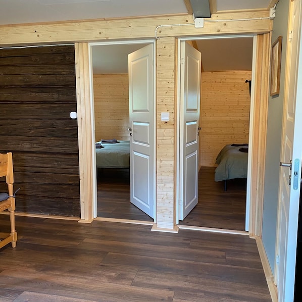Una habitación con paredes y suelos de madera, con dos puertas abiertas que dan a otra habitación con una cama y una silla.