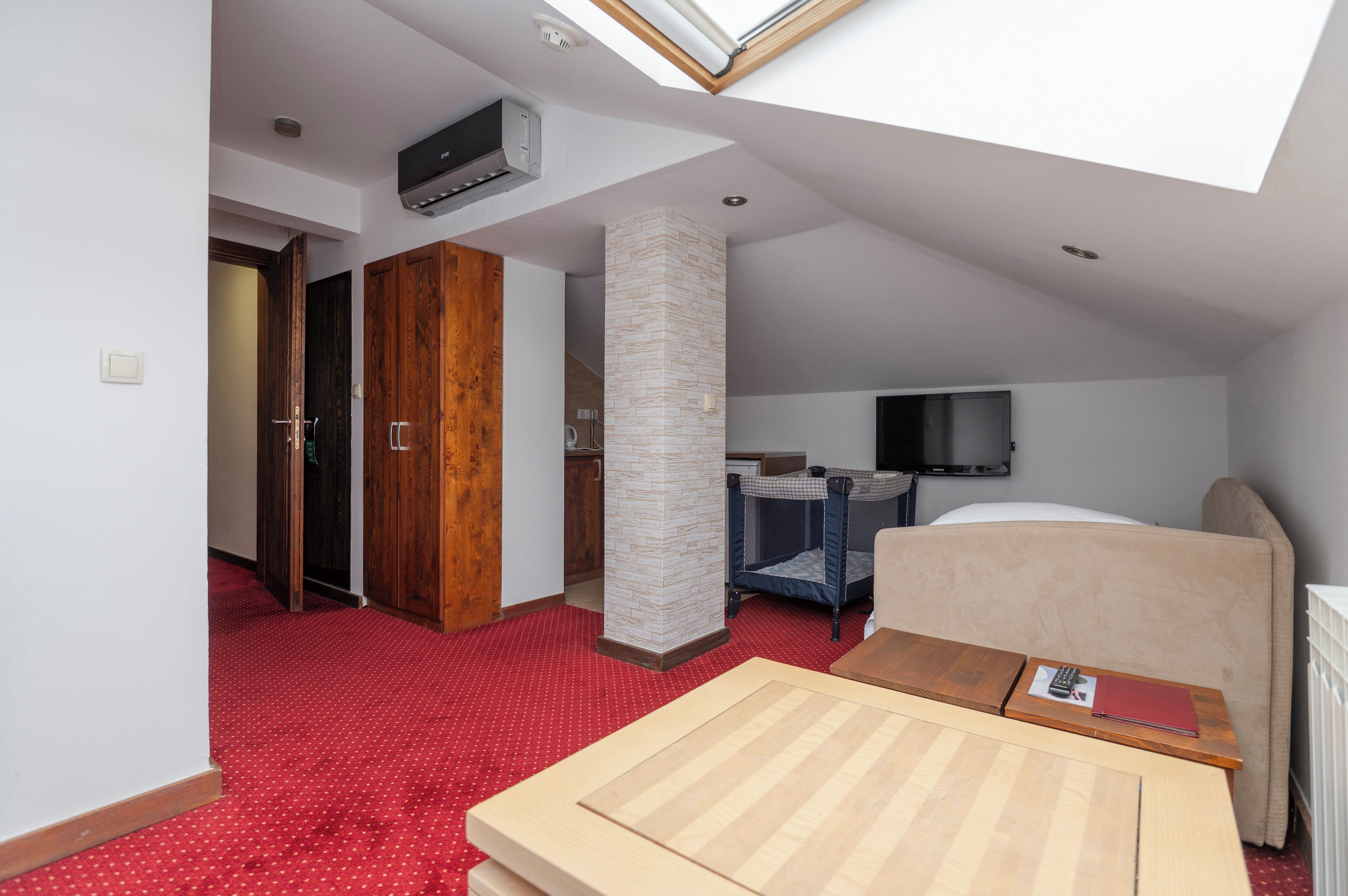Quarto de hotel com claraboia, uma cama, escrivaninha, cadeira, televisão e guarda-roupa.