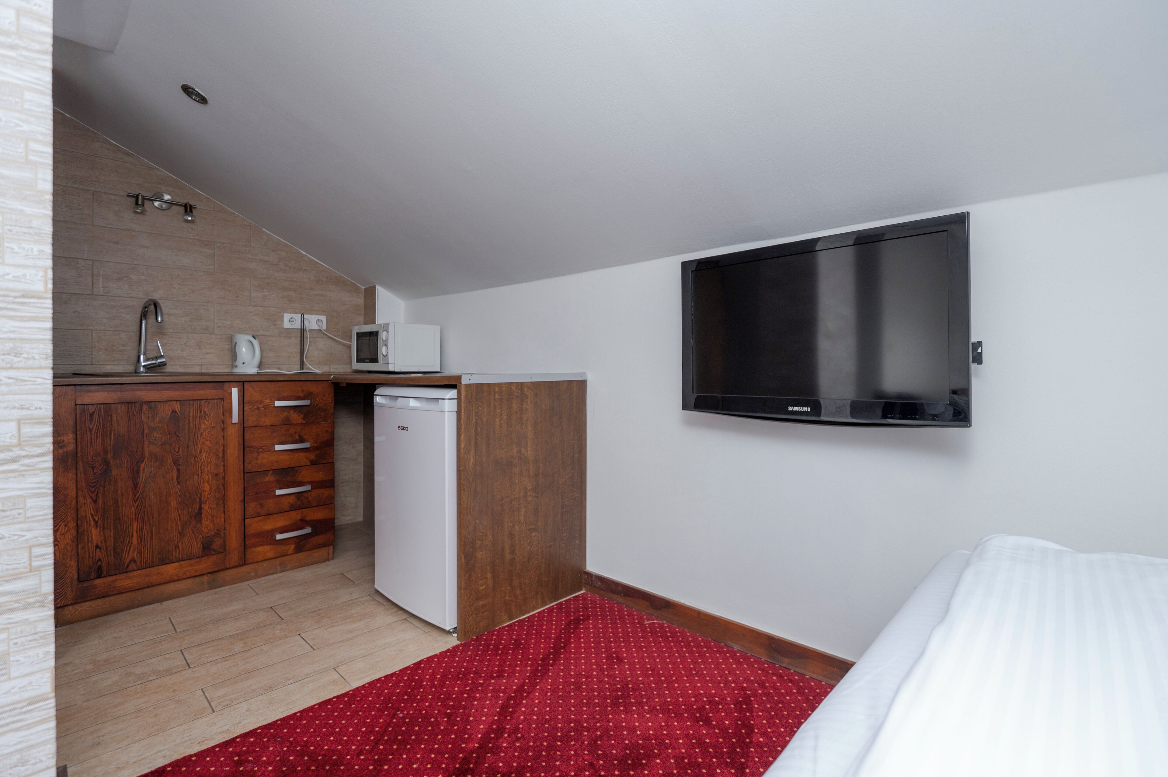 Quarto de hotel compacto com cozinha compacta, televisão de tela plana, uma cama e um tapete vermelho estampado.