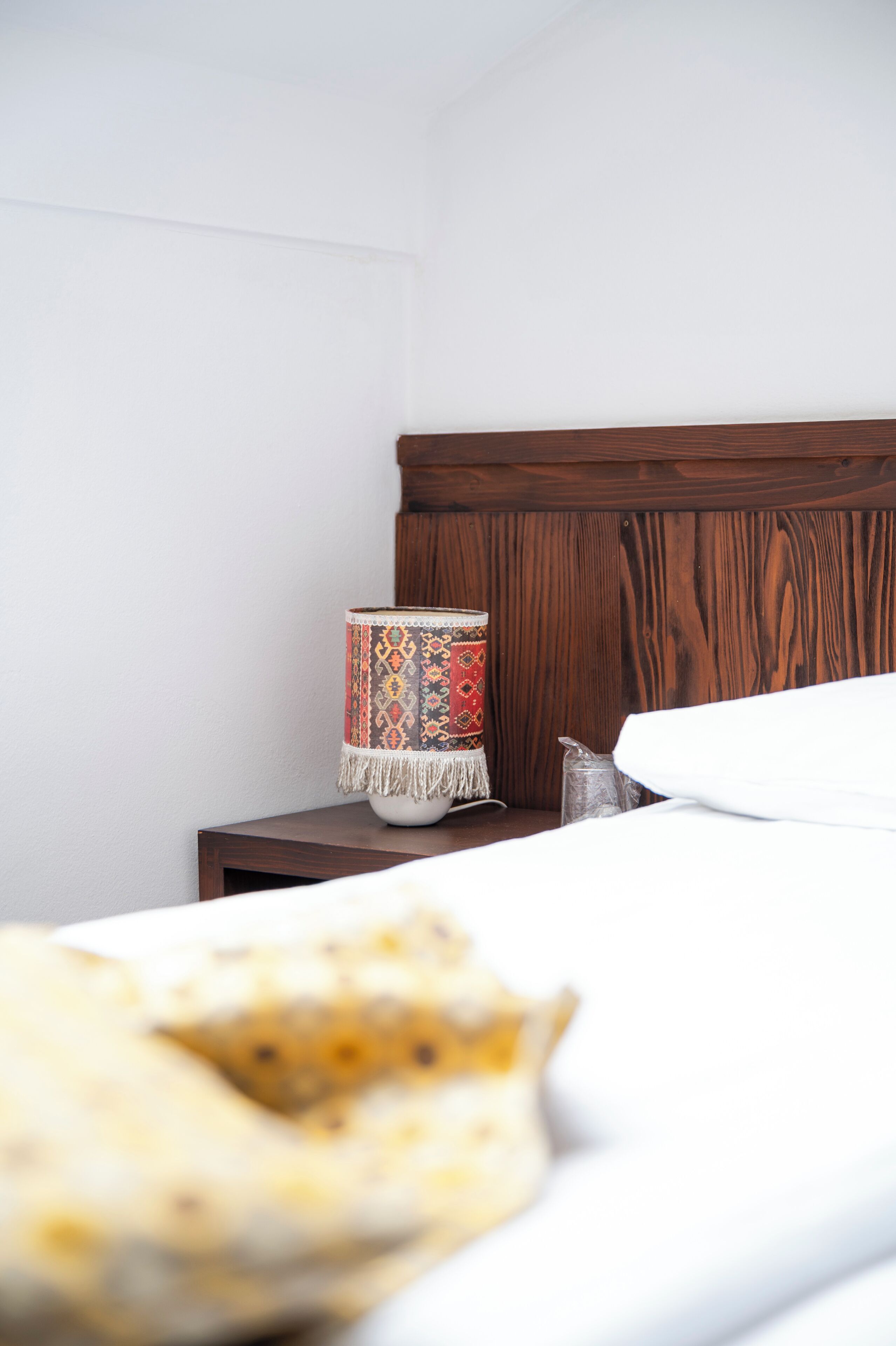 Een net opgemaakt bed met een witte dekbedovertrek en een kussen, een bedlamp met een lampenkap met patroon, en een houten nachtkastje.