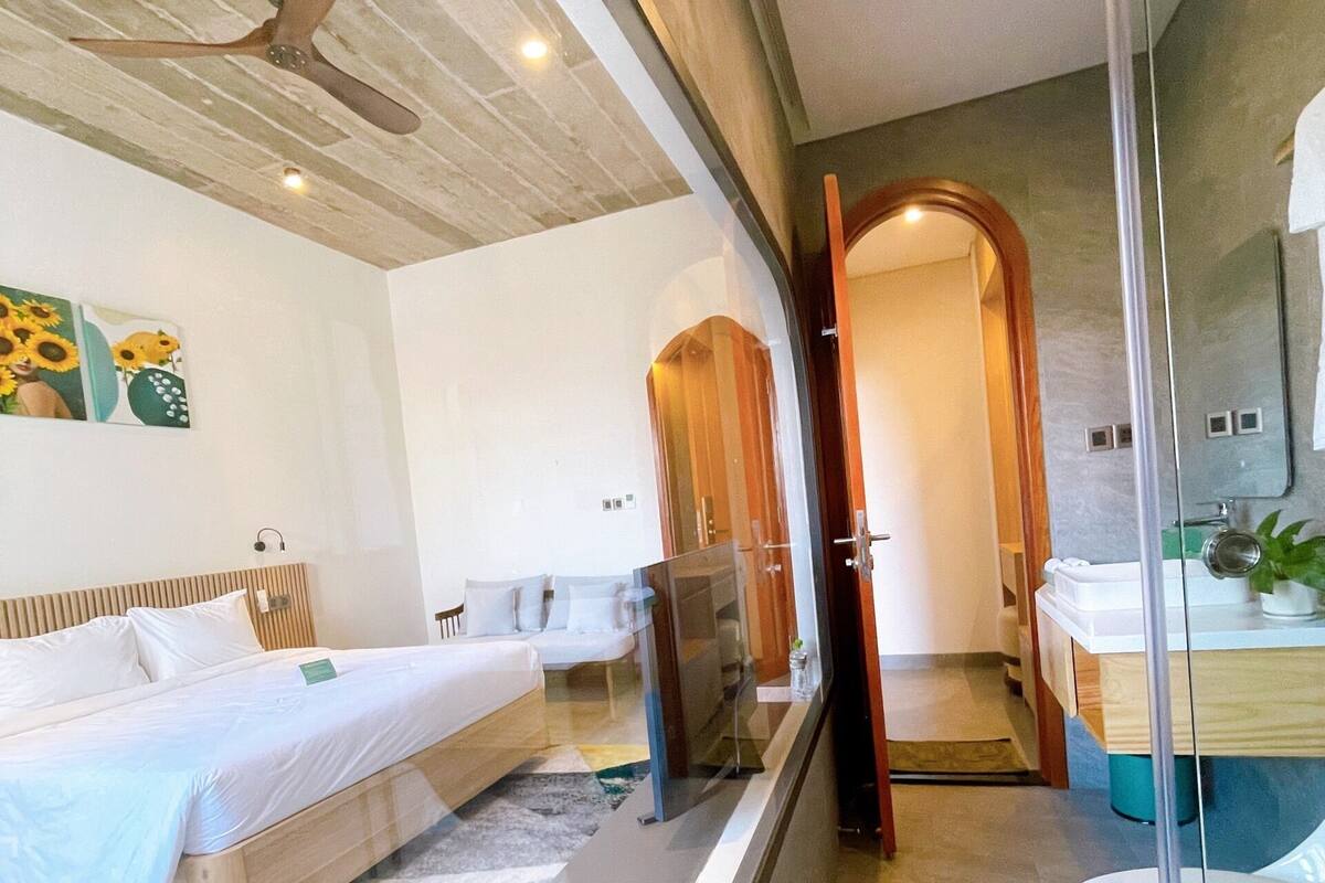 A modern hotel room with a bed, a ceiling fan, a bathroom with a toilet and sink, and a glass partition.