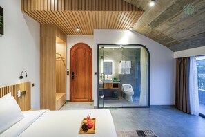 Une chambre moderne avec une tête de lit en bois, un lit agrémenté d’un plateau de pommes de pin, une porte en bois et une salle de bain fermée par des parois de verre, équipée de toilettes et d’un lavabo.