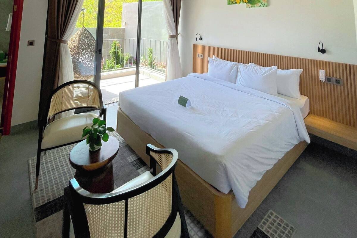 A hotel room with a bed, a ceiling fan, a table with a plant, a chair, and a view of the outdoors through a large window.