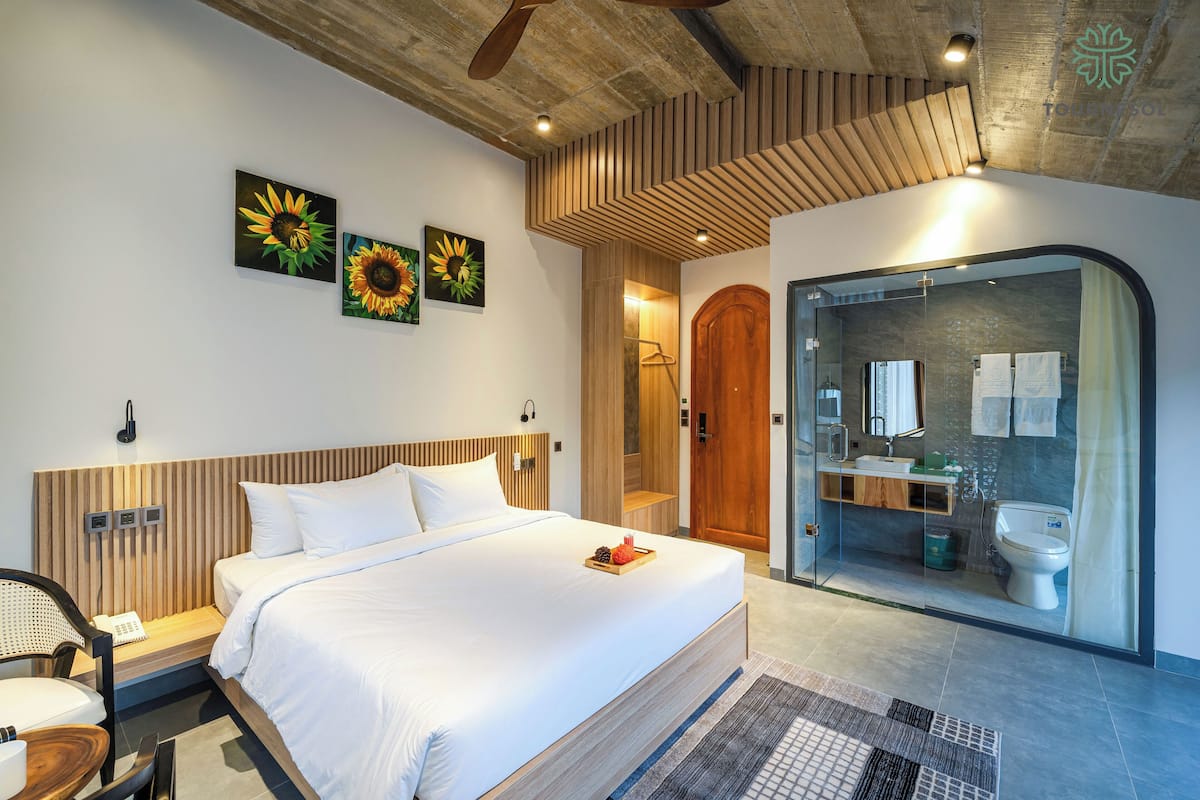A modern hotel room with a large bed, a wooden headboard, a glass shower enclosure, and a small bathroom.