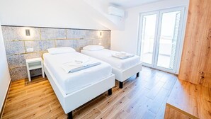A modern bedroom with two beds, a small bedside table, a tiled wall, and a large window. - Resort Trcol (Novalja)