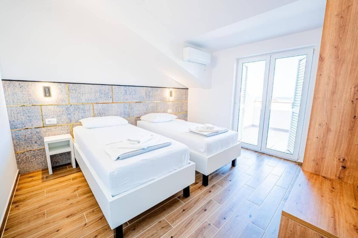 Een moderne slaapkamer met twee bedden, een klein nachtkastje, een betegelde muur en een groot raam.