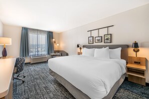 A hotel room with a large bed, a desk, a chair, a desk lamp, a bedside table, a wall-mounted lamp, and a window with curtains. - Sleep Inn & Suites Harrisburg Airport - Hershey South (Middletown)