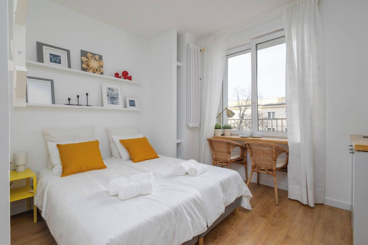 A neatly arranged bedroom with a bed, a desk, and a chair. There are shelves with framed pictures and decorative items.