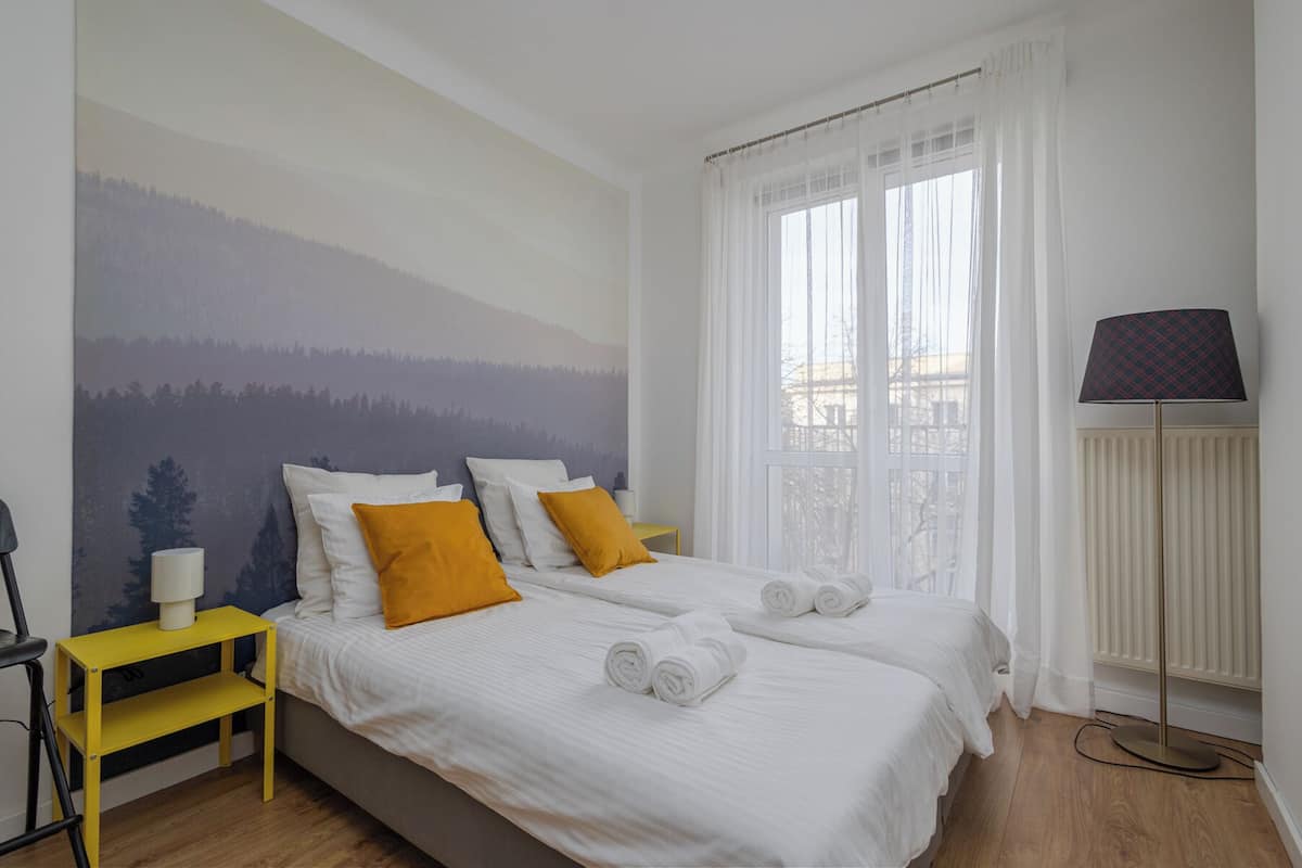 A hotel room with two beds, white bedding, yellow pillows, a mountain mural, a window with sheer curtains, a lamp, and a radiator.