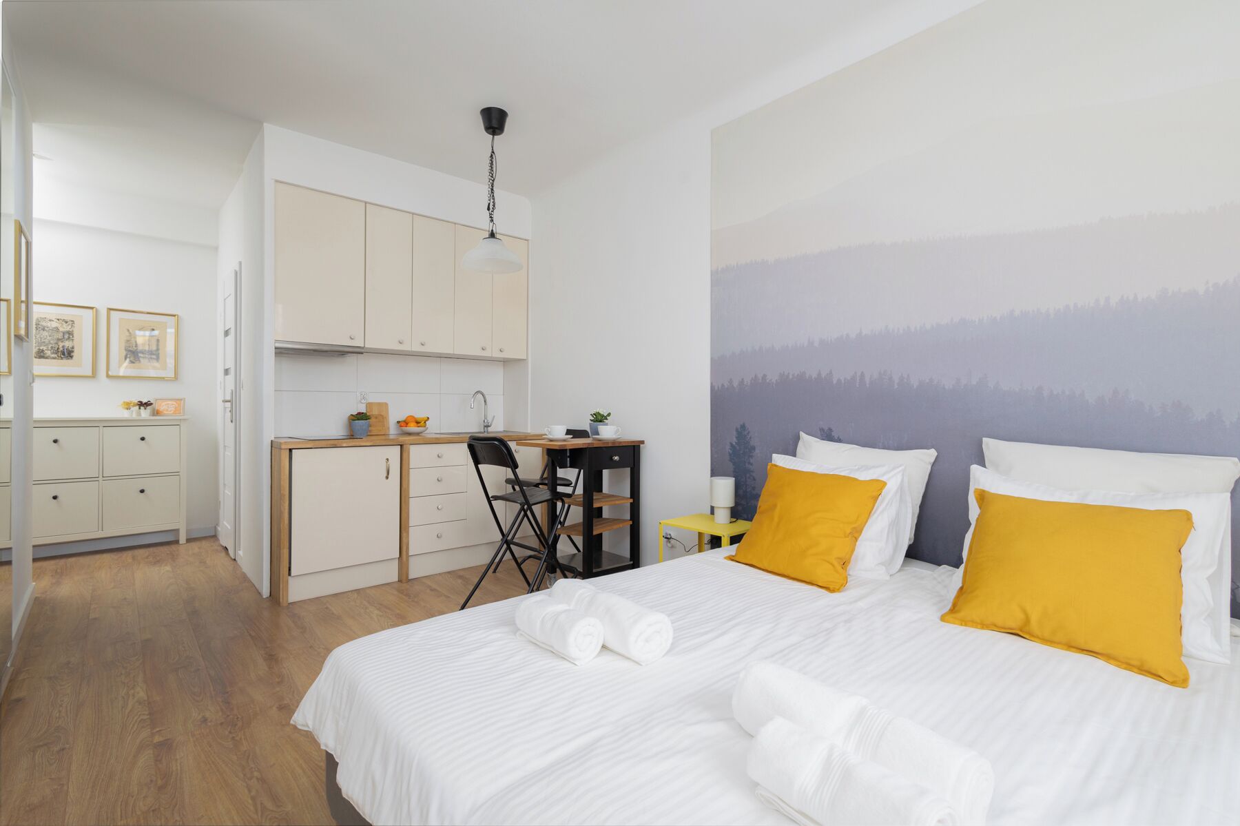 Un dormitorio con una cama, dos almohadas amarillas, un escritorio con una silla, una mesita de noche con una lámpara y una zona de cocina con armarios y un fregadero.