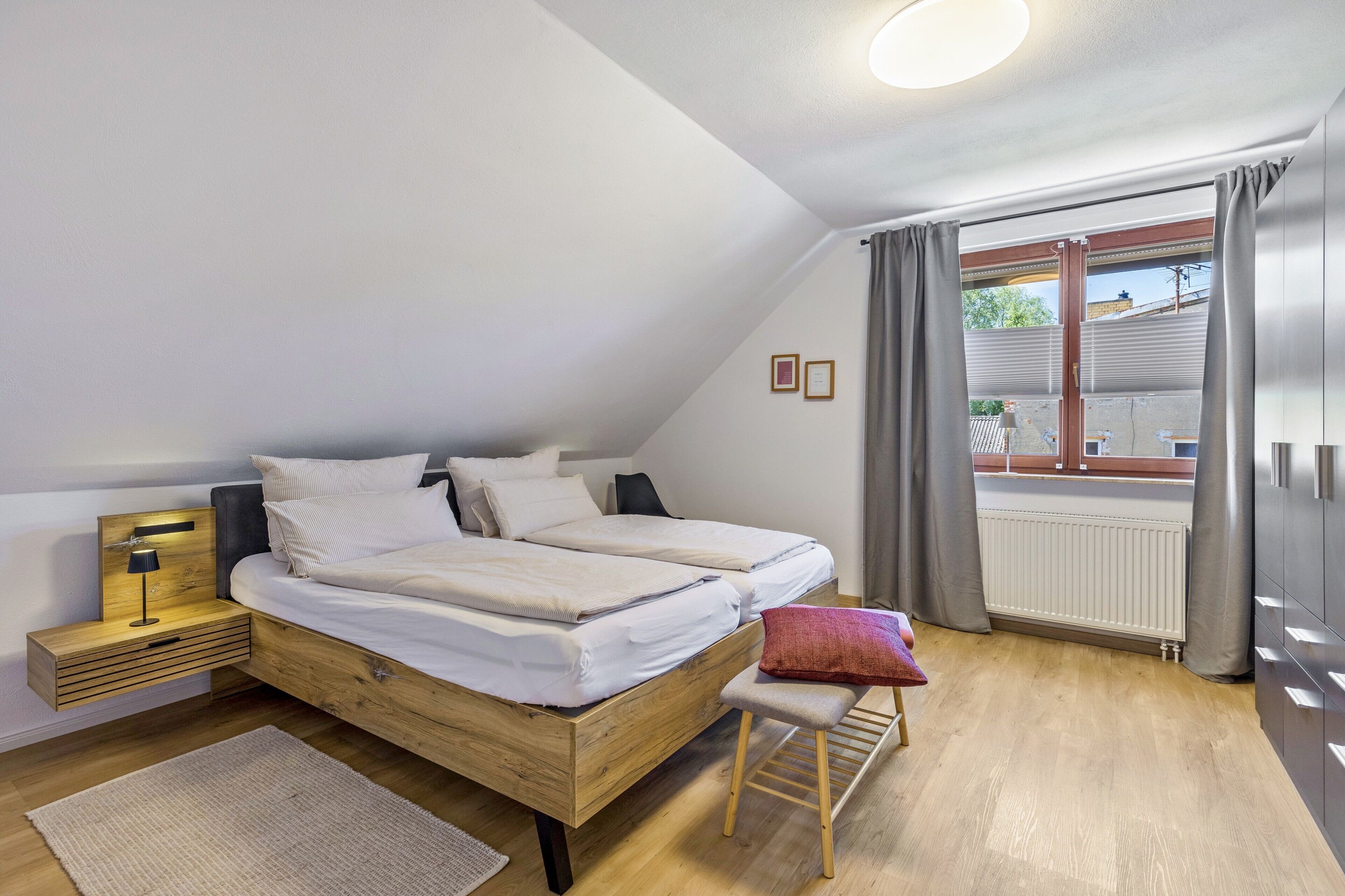 Ein Schlafzimmer mit einem hölzernen Bett, einem Nachttisch, einer Bank und Blick ins Freie.