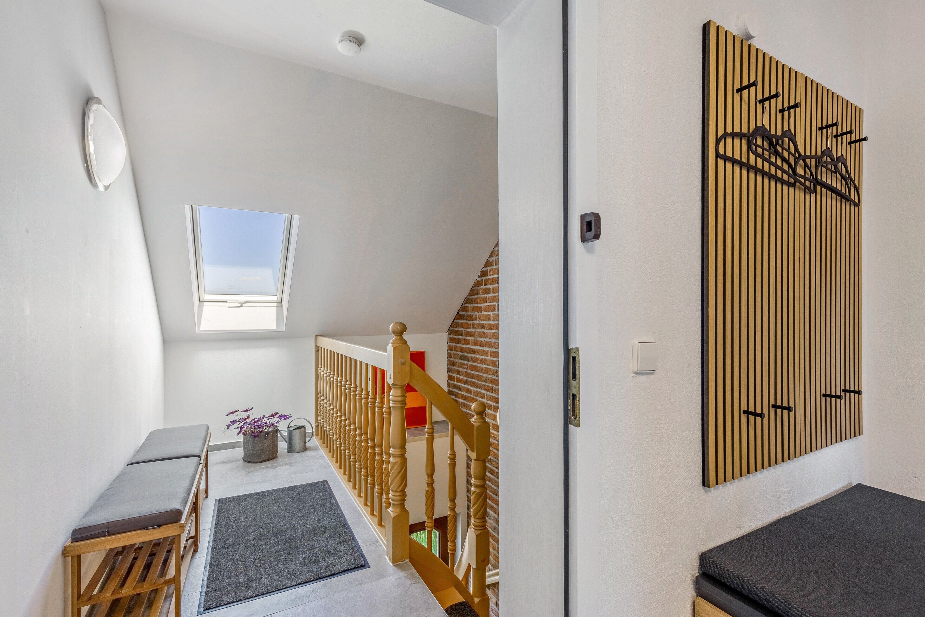Un couloir avec un banc en bois, une petite table, une plante en pot, une fenêtre et une œuvre d’art accrochée au mur.