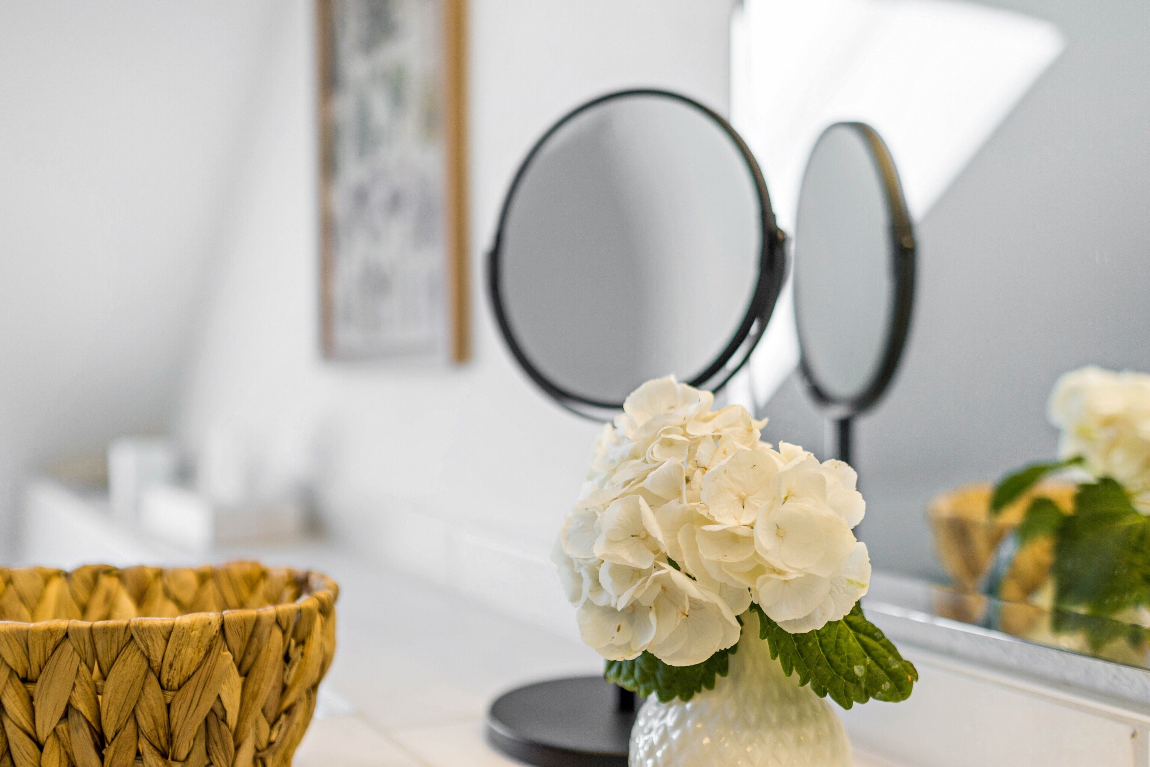 Un tocador de baño con un jarrón blanco lleno de flores, una cesta tejida y dos espejos redondos.